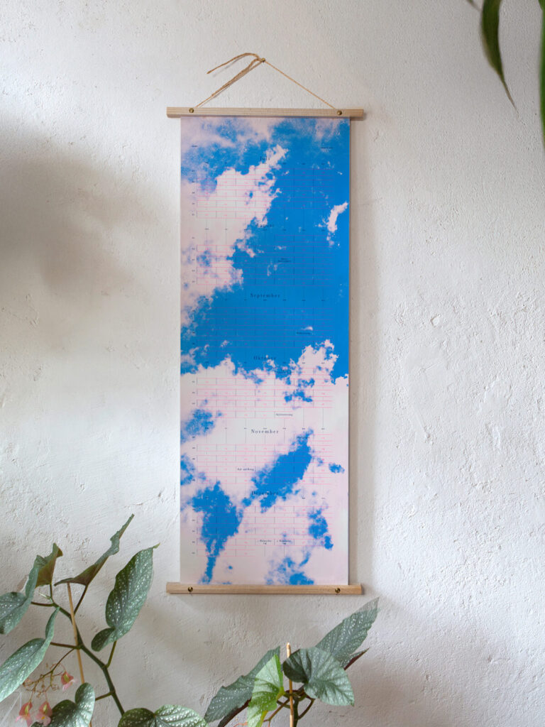 Sky High (Special Edition): Hochwertiger, limitierter und handgemacht Siebdruck-Designkalender 2025 von days & dots an einer weißen Wand hängend mit Pflanzen
